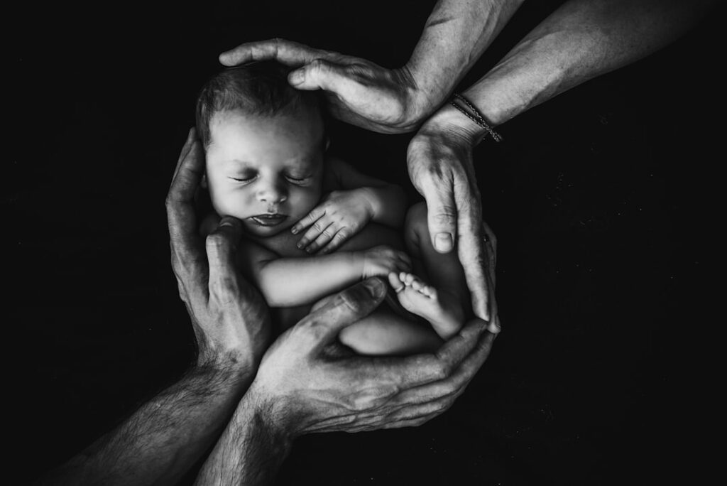 Black and white photo. An infant being held byt he hands of both a woman and a man. Credit: Isaac Quesada on UnSplash